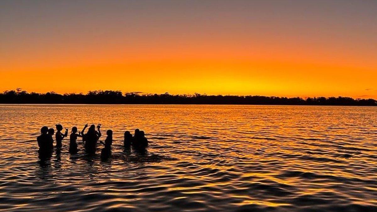 Students stand in the ocean against a bright orange sunset.